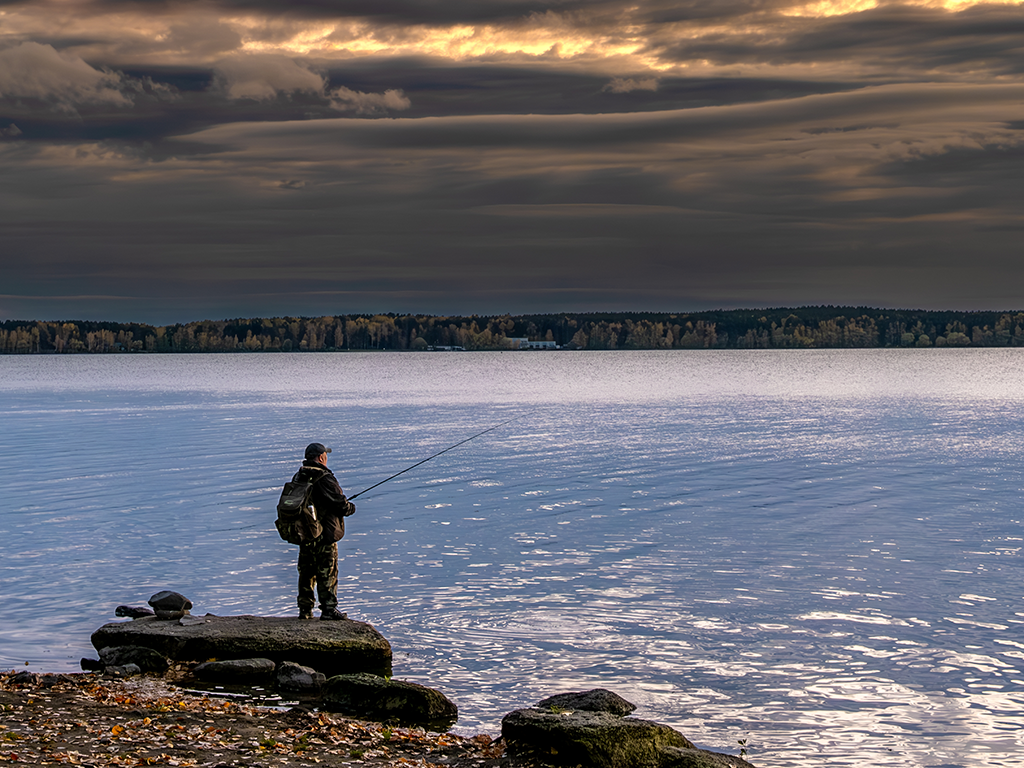 A lone angler fishes from a rocky shoreline under heavy cloud.