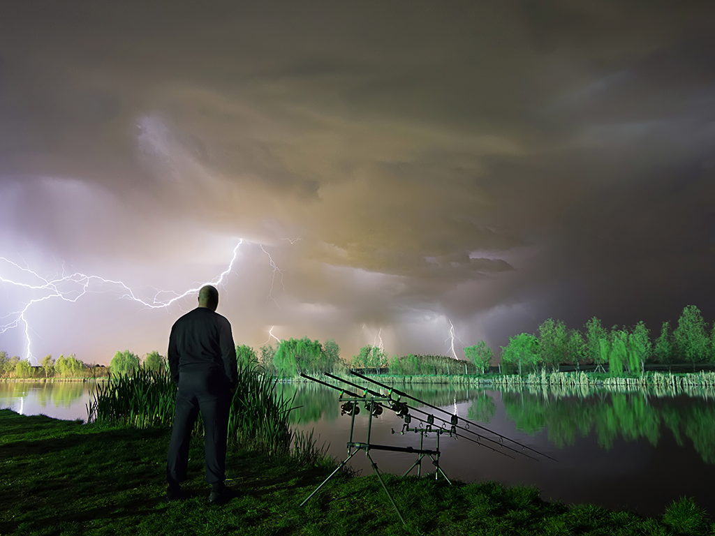 A view from behind of a man next to some rods set up for shore fishing, waiting for a thunderstorm to pass with lightning visible in the sky above him.
