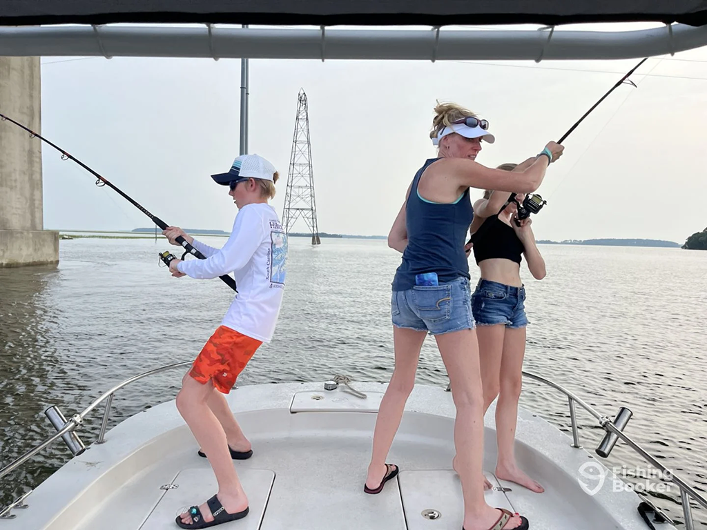 Two teenagers and their mother casting a fishing line from the hull of a fishing boat near some rigs in shallow water.