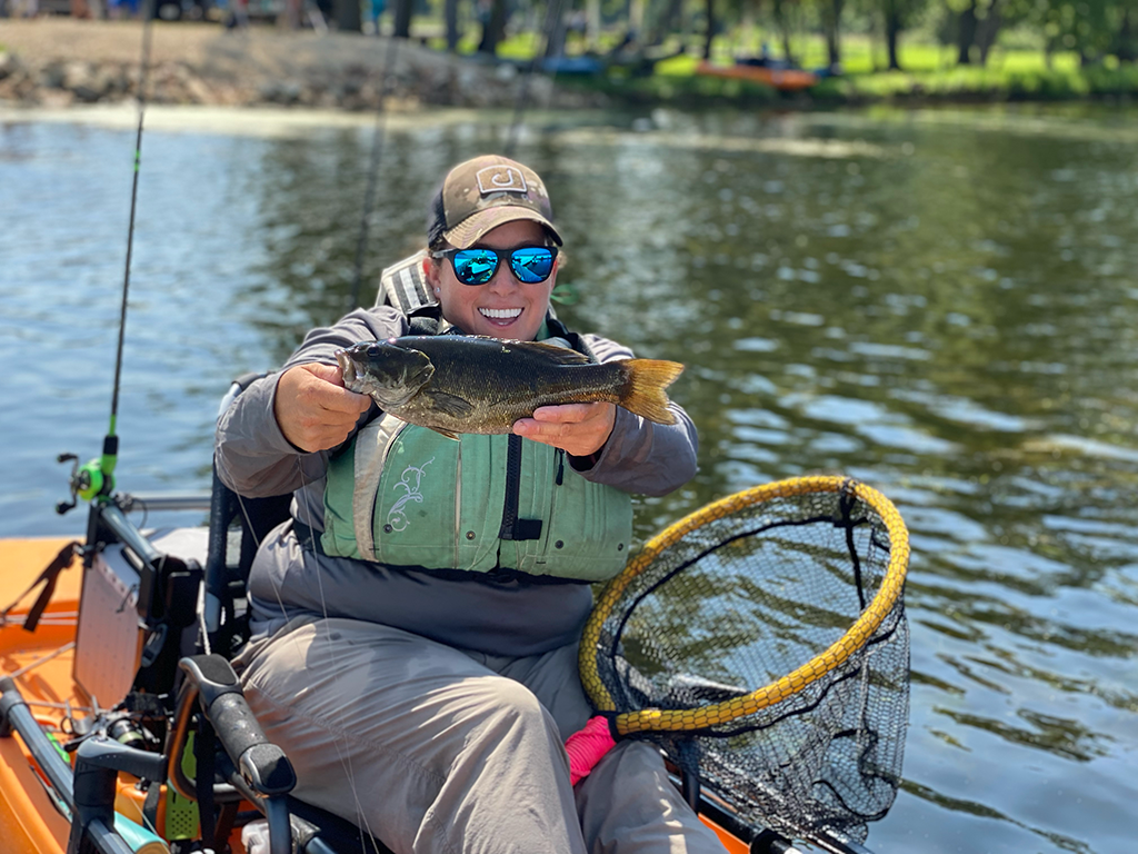 A woman sitting on a kayak and holding a Smallmouth Bass to the camera to one of the fishing events organized by the WFF on a sunny day.