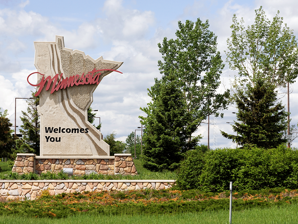 A roadsign saying Minnesota Welcomes You on the outline of the state map visible next to trees on a day with sunny intervals