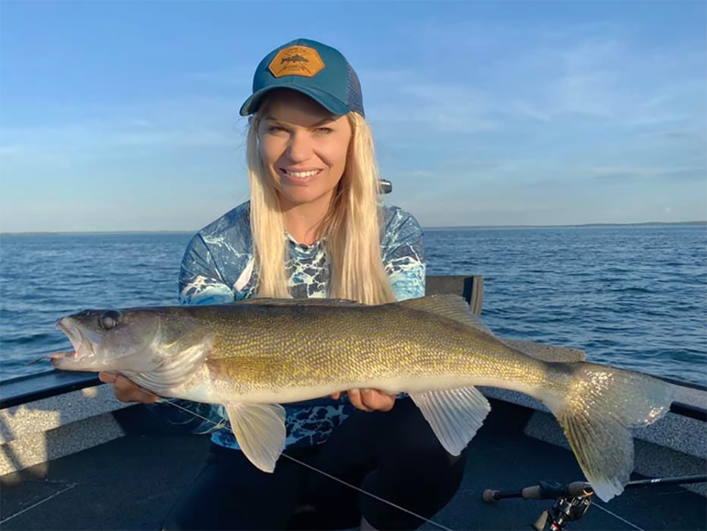 A woman in a baseball cap holding a Walleye on a lake in Minnesota on a clear day.