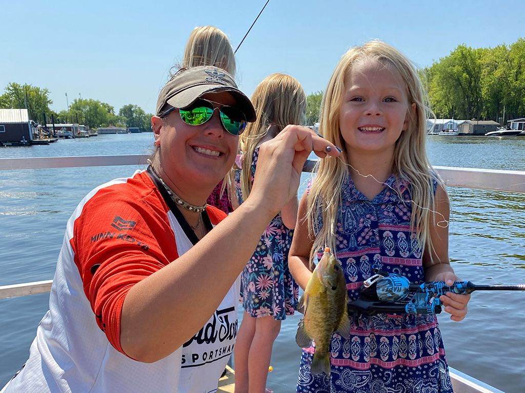 A woman and little girl crouching down on a boat and holding up a small Panfish on a sunny day.