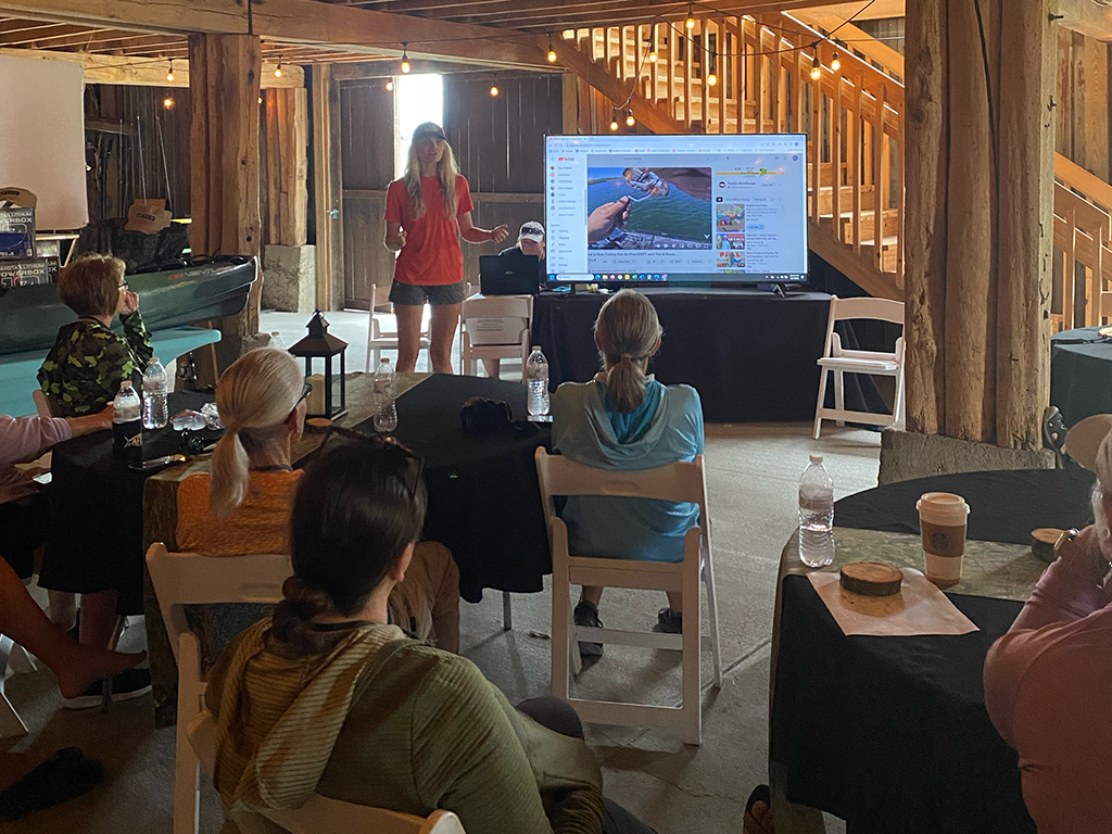 A seminar being held by the Women's Fishing Federation, with a woman in red presenting next to a big screen in front of people in a wooden cabin.