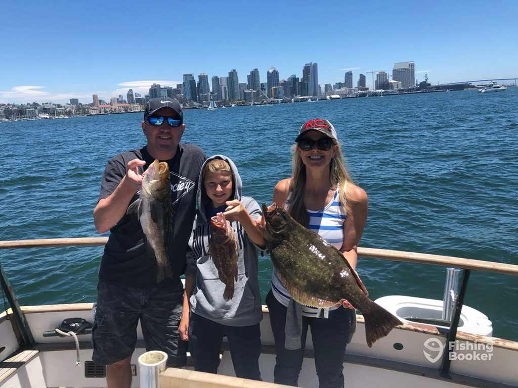 A man, woman, and child stand on a fishing boat in front of the San Diego skyline on a clear day, each holding a fish they caught on a trip.