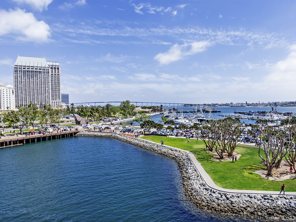 A waterfront park in San Diego Bay with green lawns and trees sits next to a marina filled with boats. A large building's visible on the left, and a bridge stretches across the background under a partly cloudy sky.