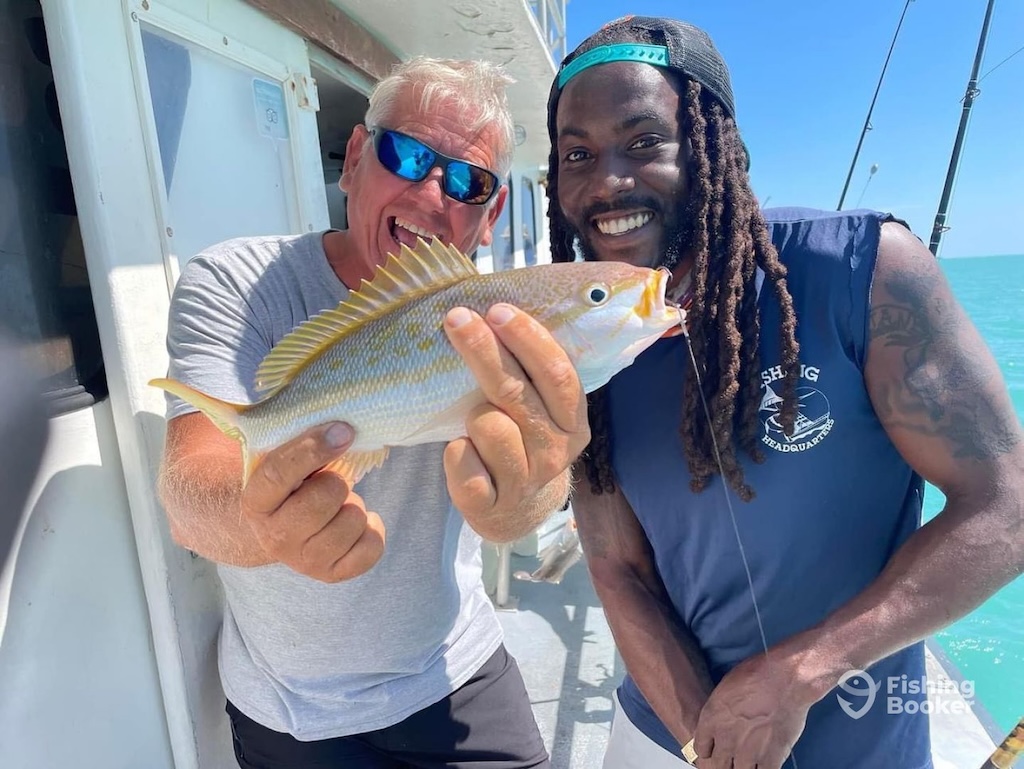 Two men are on a boat, smiling at the camera. One's holding a freshly caught Yellowtail Snapper.