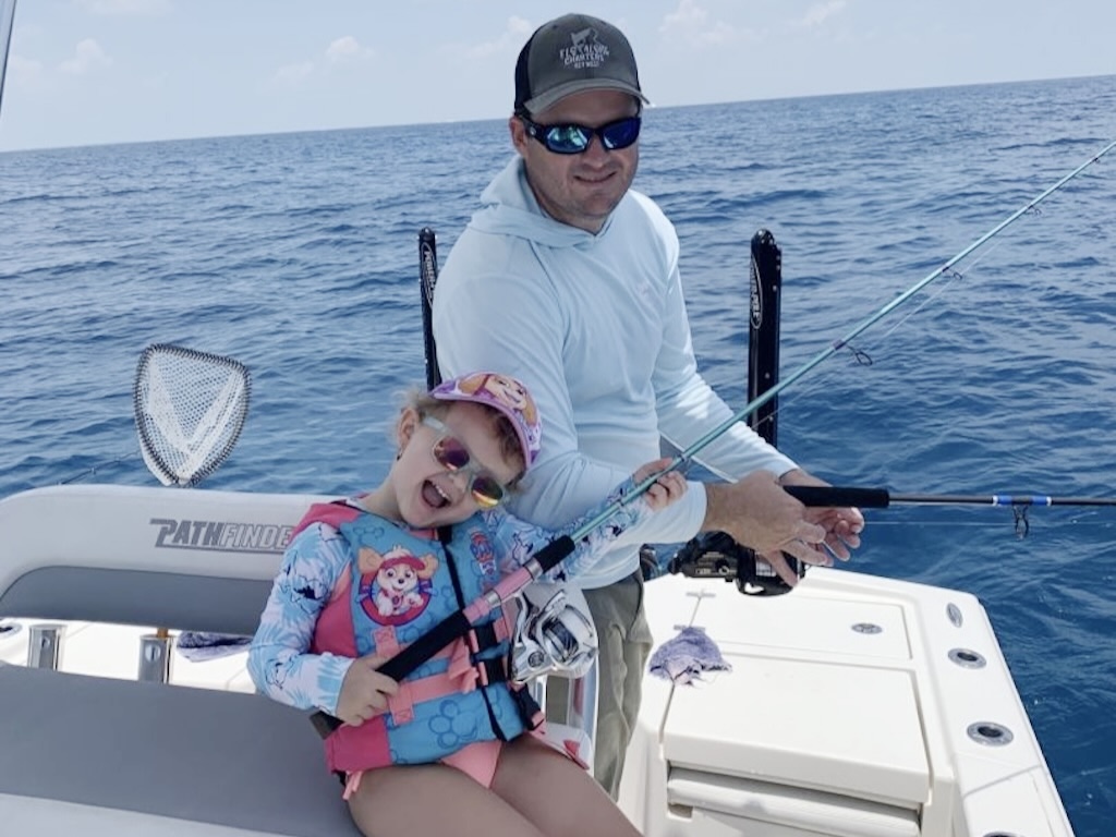 An adult and a child are Yellowtail Snapper fishing on a boat in open water. The smiling child, who's wearing a life jacket and cap, proudly holds a fishing rod.