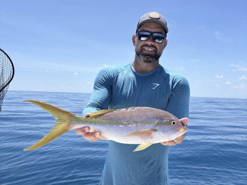 A man in sunglasses and a cap holds a freshly caught Yellowtail Snapper aboard a boat, enjoying a perfect day of fishing under clear blue skies.
