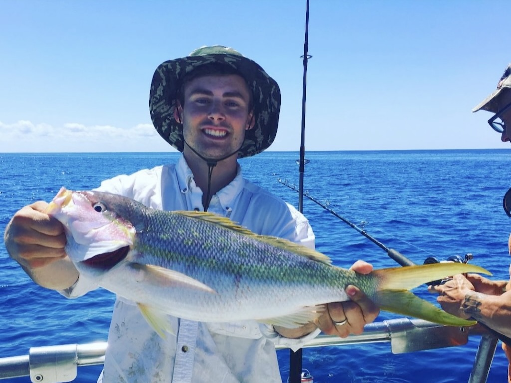 A person wearing a bucket hat and white shirt is holding a large Yellowtail Snapper on a boat, with fishing rods and the ocean visible in the background.
