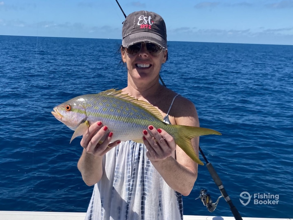 A person on a boat holds a Yellowtail Snapper in both hands, enjoying an exciting Yellowtail Snapper fishing trip with the ocean and a fishing rod in the background.