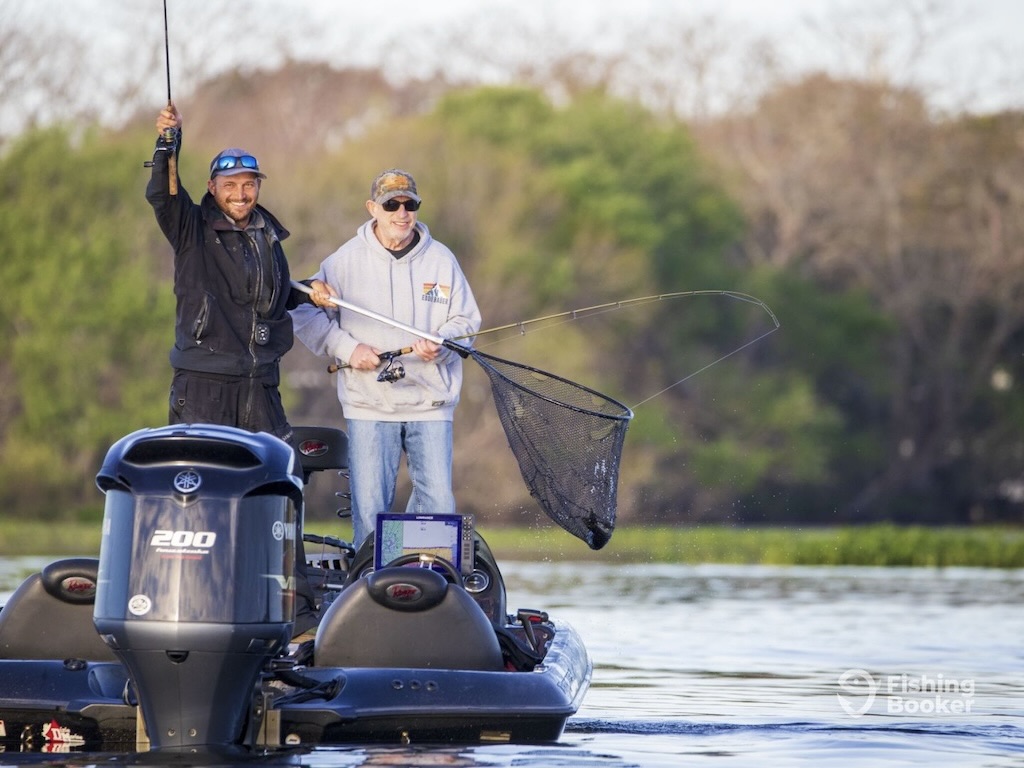 Two men on a boat, one holding a fishing rod and another with a net in his hand, celebrarte as they hook a Chain Pickerel fish aboard.