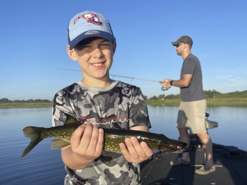 A young child holds a small Chain Pickerel on a boat in front of a man who's casting into calm lake waters on a clear day.