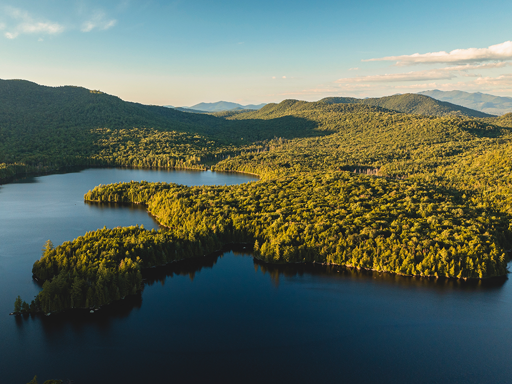 An aerial view of a lake in the Adirondack Mountains in New York with a green peninsula jutting out into the lake on a clear afternoon.