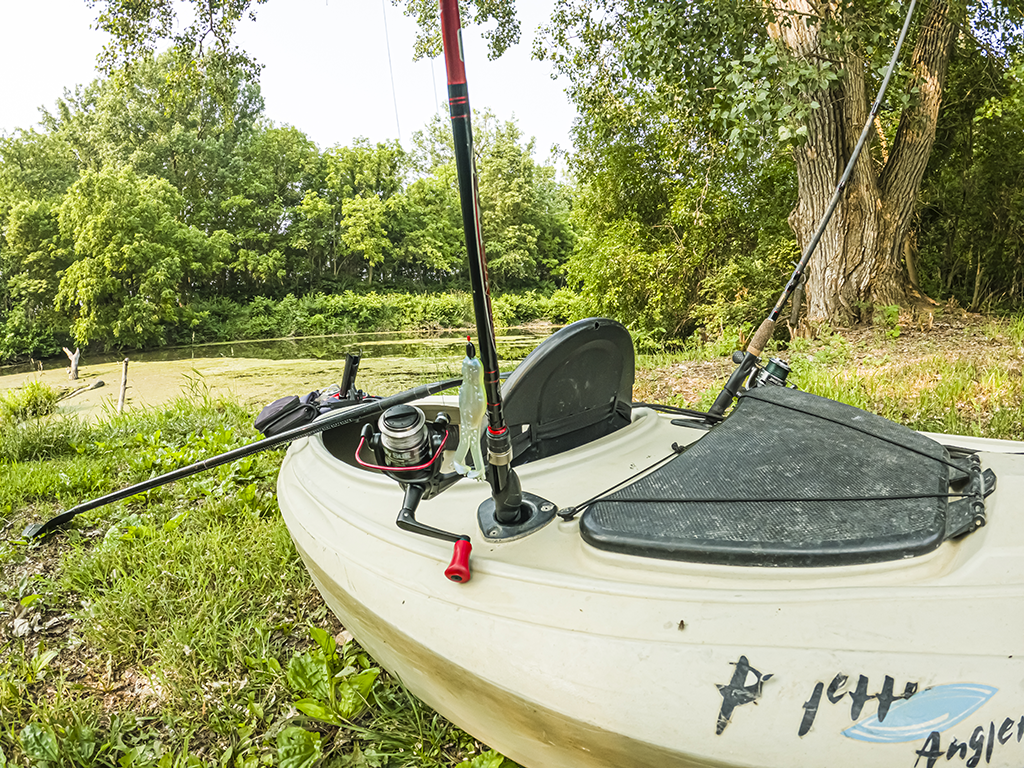 A kayak laying on some grass and under some trees, fitted out for fishing with a rod visible in a rod holder.