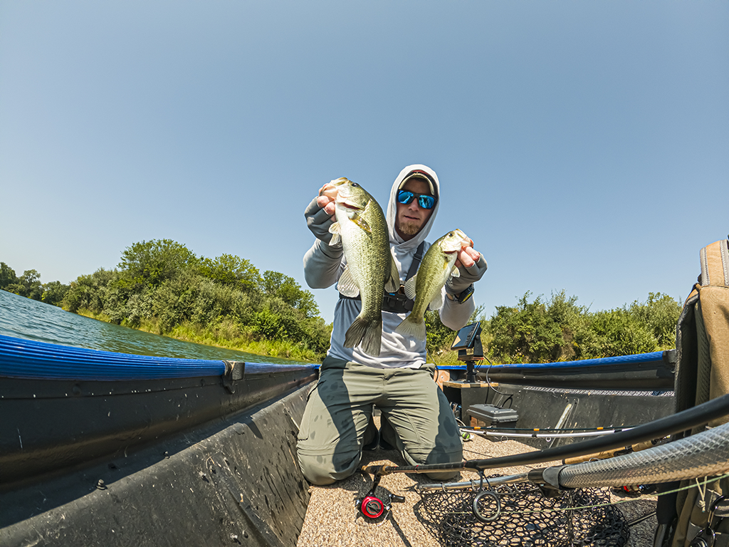 A man in a hooded jumper and sunglasses on a narrow boat holding up two Bass fish on a clear day