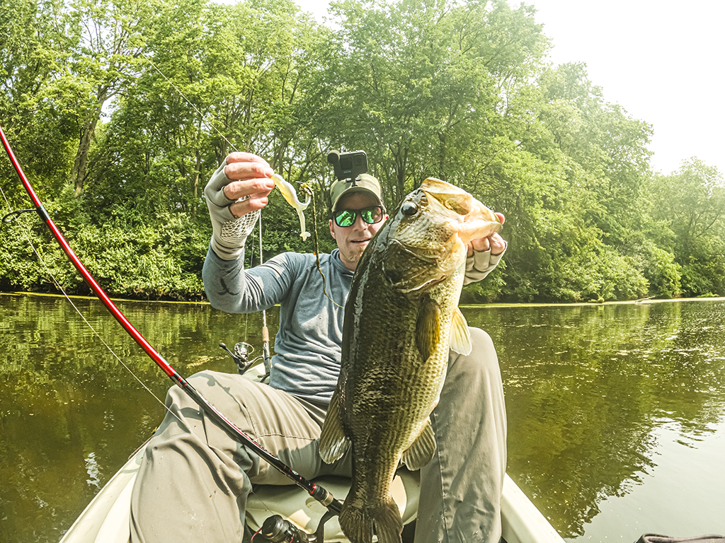 A man in a hat and with a GoPro on it holds up a Smallmouth Bass in one hand and the lure that caught it in another while sitting on a kayak.