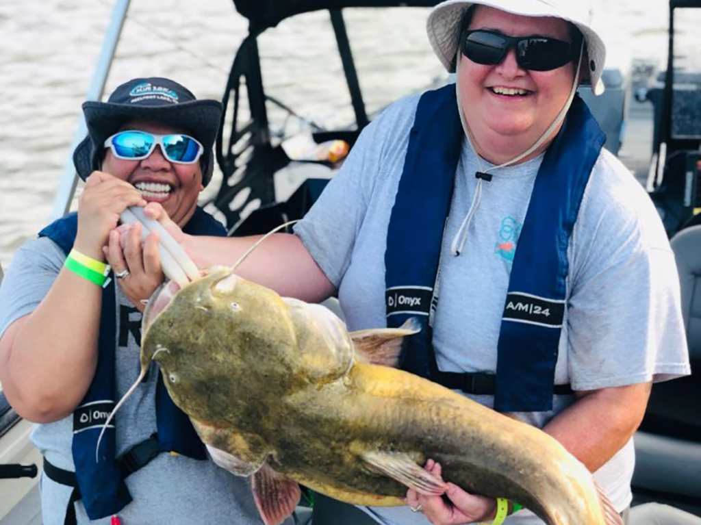 Two people on a boat holding up a large, yellow Flathead Catfish, while wearing hats, sunglasses, and safety jackets. 
