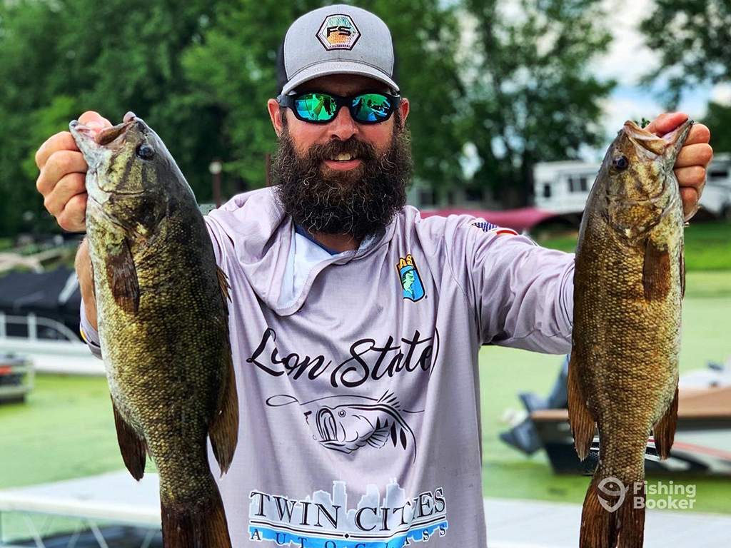 A man in sunglasses and a baseball cap holding up two Bass fish after a successful day fishing in Lake Vermilion