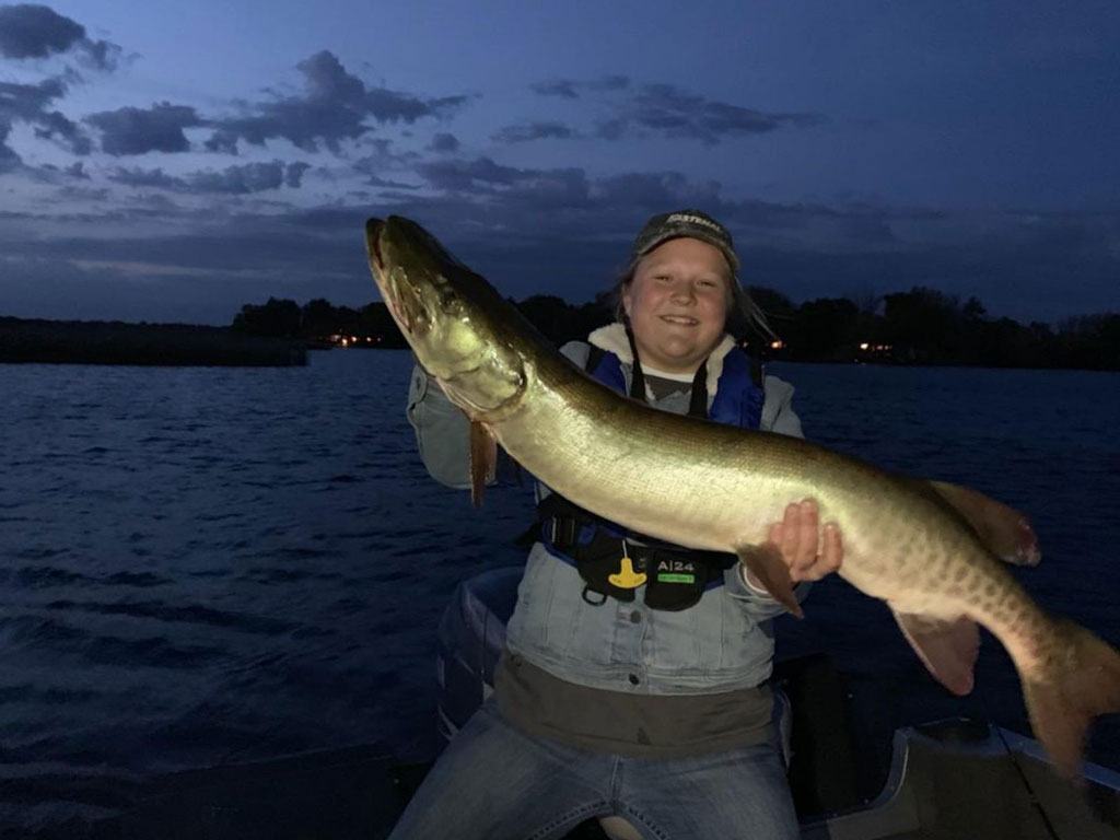 A nighttime photo of a teenager holding a large Musky next to a lake in Minnesota.