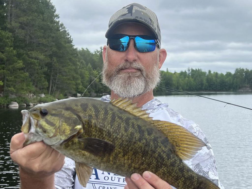 A man in sunglasses and a baseball cap holds up a medium-sized Bass next to a lake on a cloudy day.