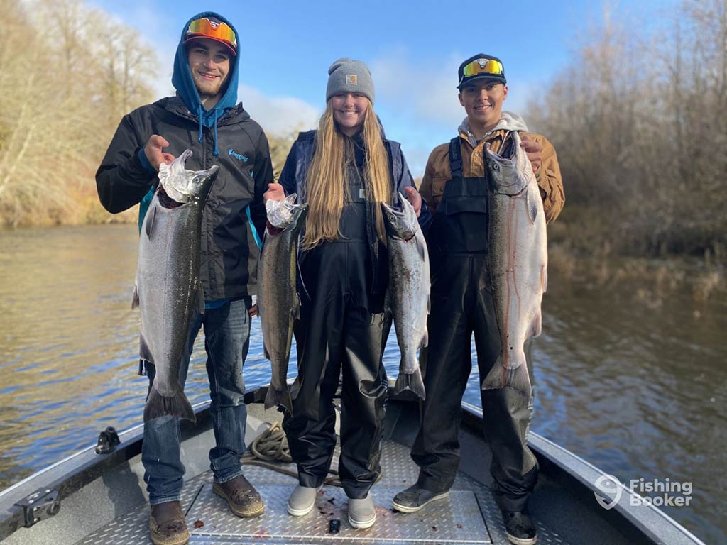 Three people standing on a shallow-water fishing boat and holding up a few Salmon each with a river setting visible around them.