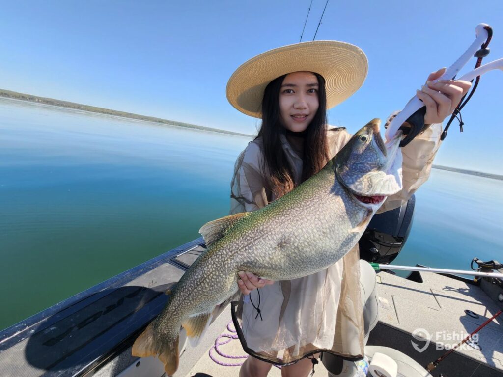 A woman in a wide-brimmed hat’s holding a large Lake Trout on a boat, with calm blue water and a clear sky in the background.