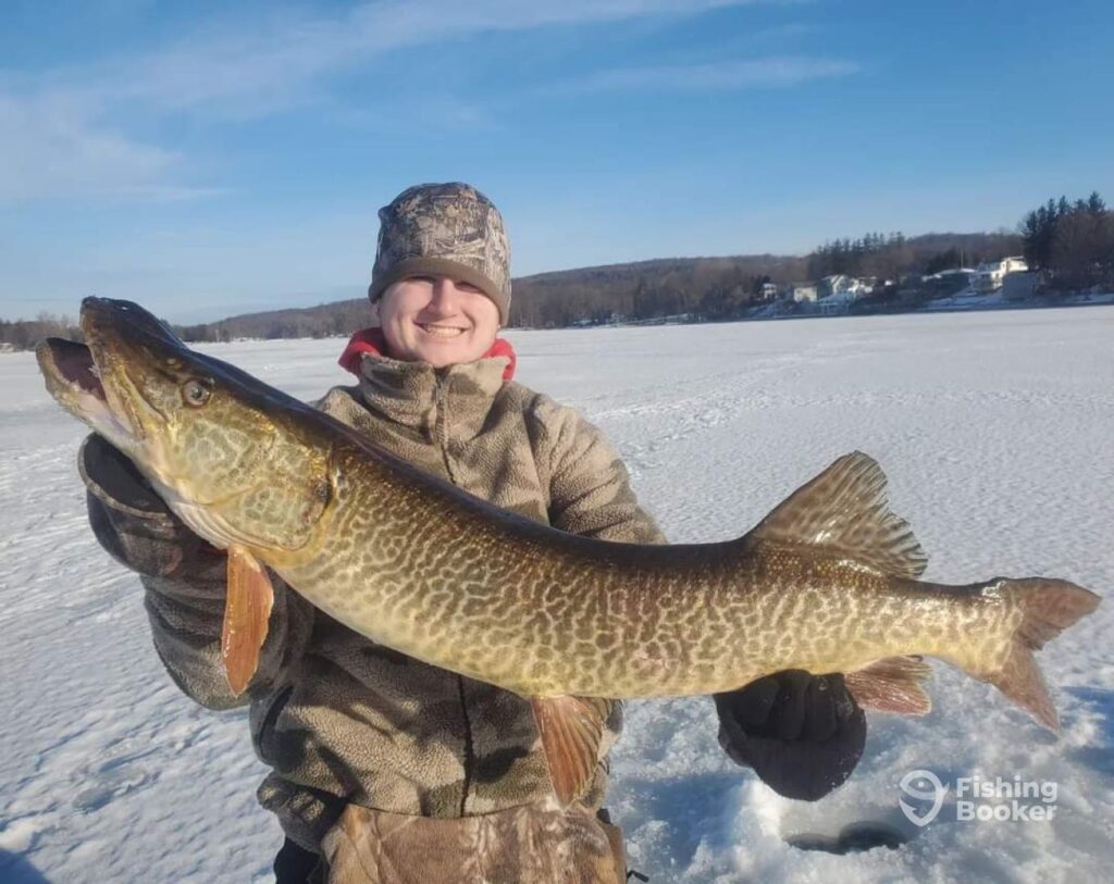 A person in winter clothing’s holding a large Musky with beautiful patterned scales. They’re standing on a frozen lake in New York's Finger Lakes, surrounded by snow and trees in the background.