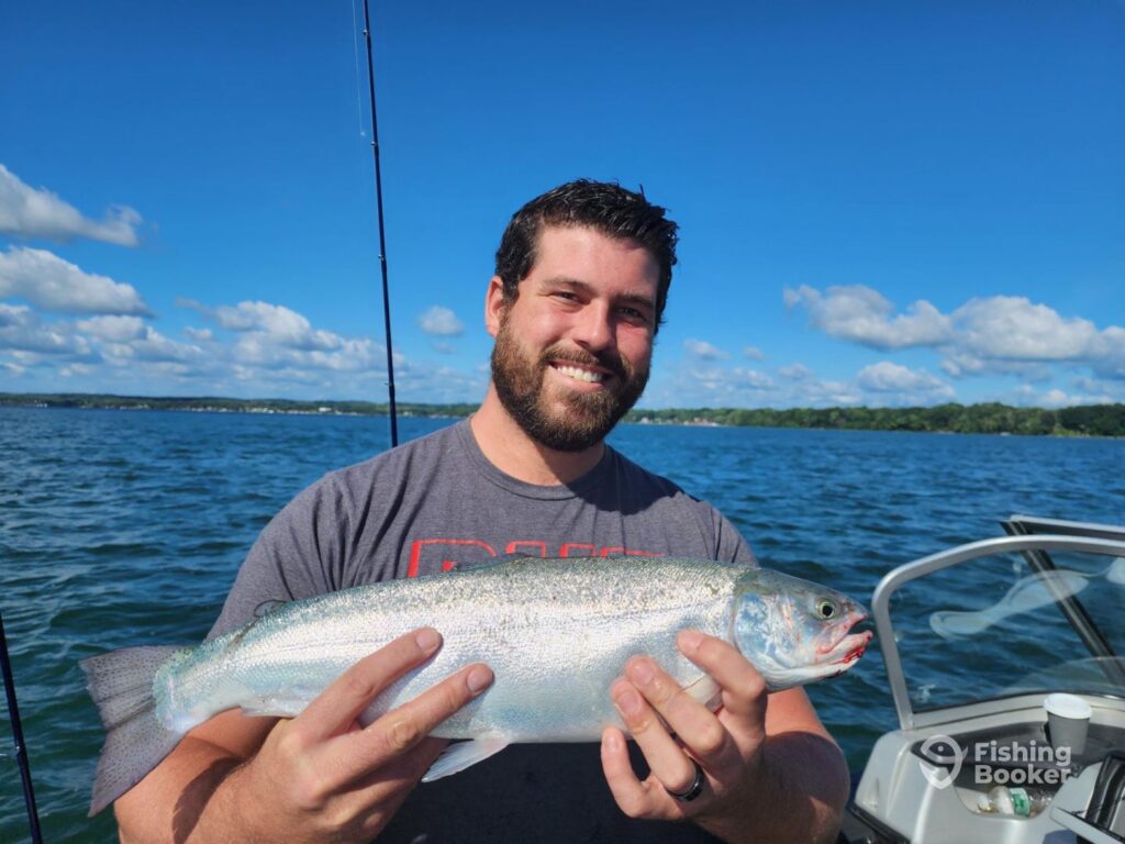 A man standing on a boat, holding a freshly caught Salmon with both hands on the open waters of one of New York's Finger Lakes on a clear day.