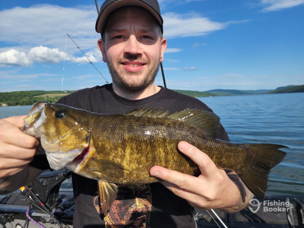 A man holding a large Bass on a boat, with a lake and hills behind him under a bright blue sky.