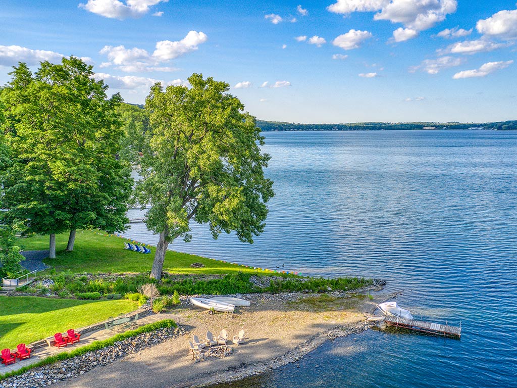 A peaceful lakeside scene in the Finger Lakes stretches before the camera. There's a lush, grassy area bordered by large Oak and Maple Trees, their leaves swaying in the gentle breeze.