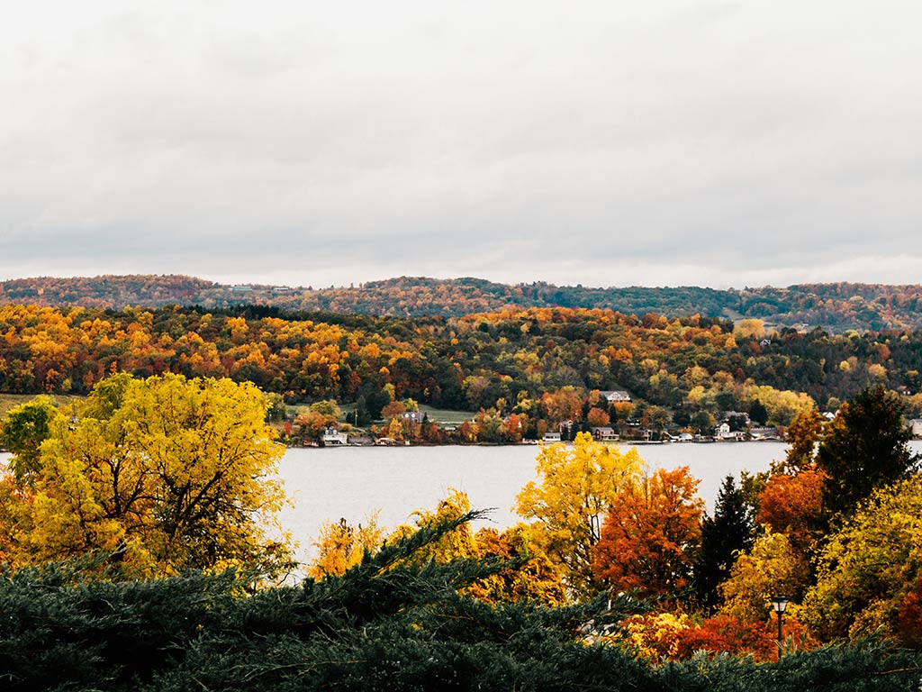 A lake is surrounded by rolling hills and trees with autumn foliage in shades of yellow, orange, and green under a cloudy sky.