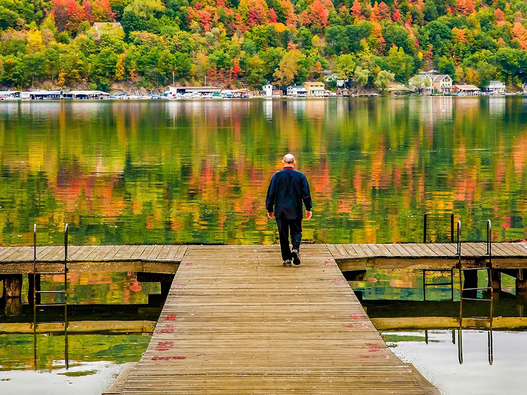 A person walking alone on a wooden dock toward a calm body of water in the Finger Lakes, where colorful autumn trees and houses are reflected in the water.