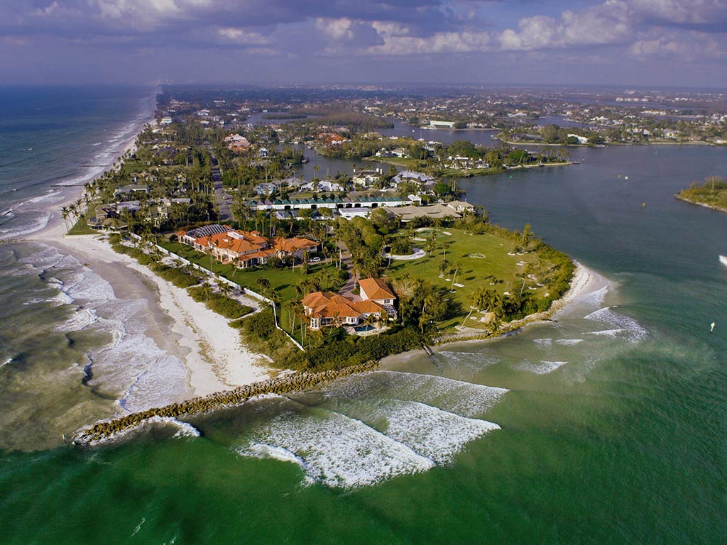 From above, you see a stunning coastal estate with large houses lined along the shoreline in Naples, FL. Green lawns stretch out around each home, dotted with tall palm trees that sway gently in the ocean breeze. All around, the blue water from inlets and the open ocean hugs the land, creating a striking contrast with the vibrant greenery. Under a partly cloudy sky, sunlight gleams off both roofs and waves alike, highlighting this picturesque seaside retreat.