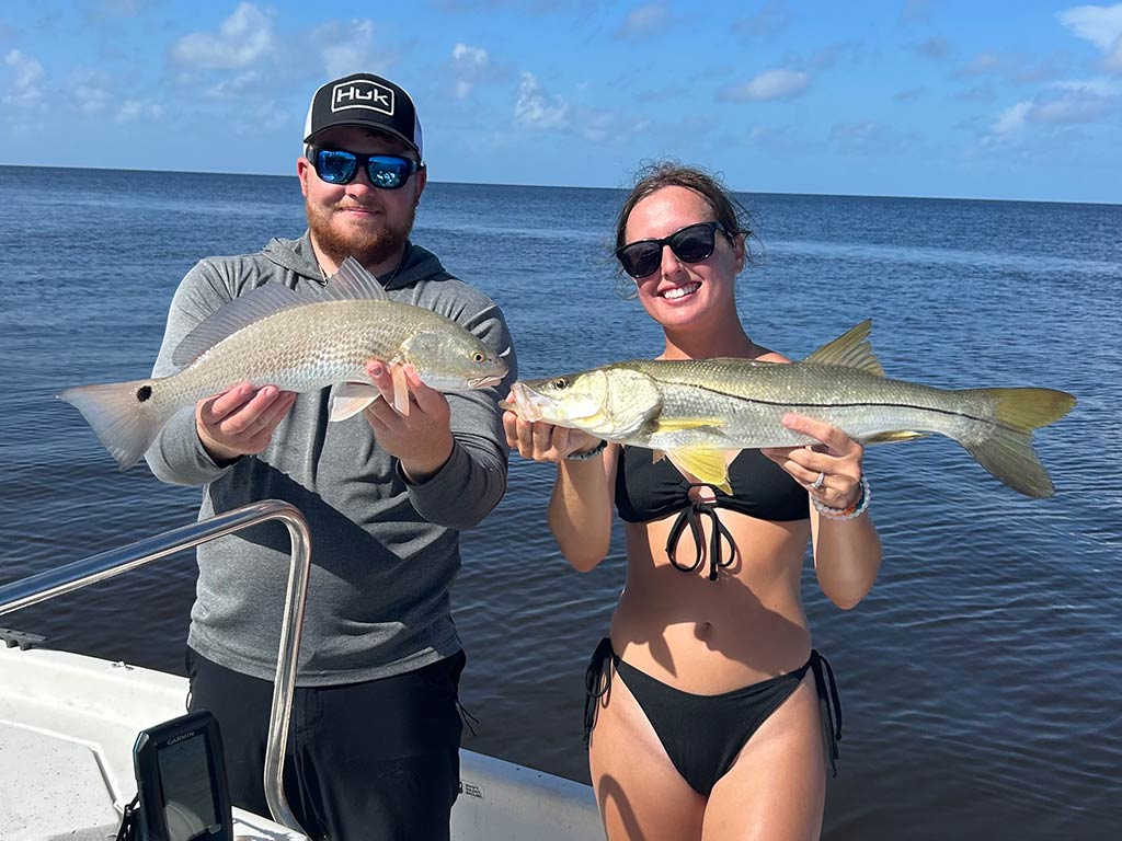 A man and woman are on a boat, fishing on a date – possibly for the first time – holding up freshly caught fish. One person is holding a Redfish, while the other is proudly displaying a Snook. Behind them, you can see water stretching out under a clear blue sky.