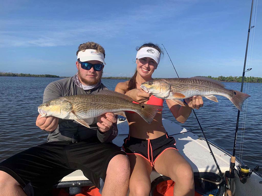 Two people are sitting on a boat on a fishing date, each holding up a large Redfish they’ve caught. Their fishing rods lean casually nearby, proof of their success. Behind them, calm water stretches out, bordered by lush trees that frame the background and add to the peaceful atmosphere of their day on the lake.