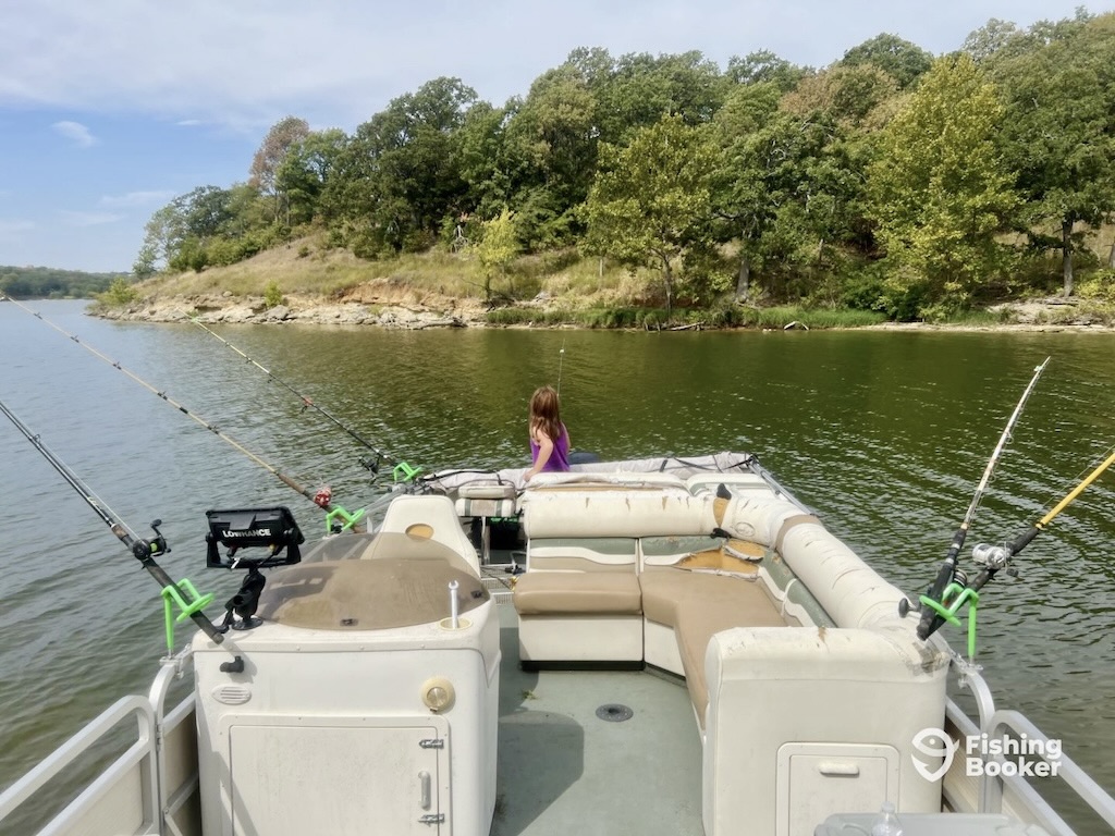 A child sitting on a bench at the back of a fishing boat, facing the lake. Fishing rods are set up along the railing, and you can see trees on the shoreline ahead.