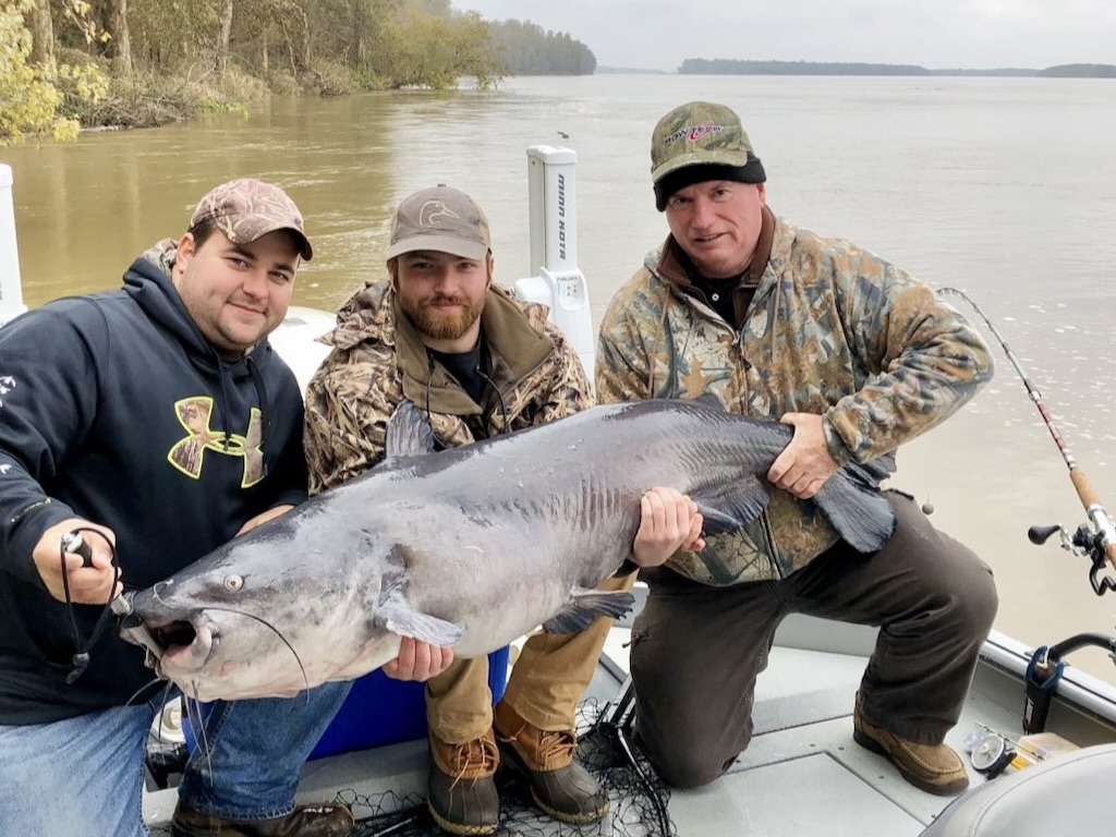 Three men on a boat are holding a large Blue Catfish they've caught, posing for a photo with fishing rods and a river in the background.