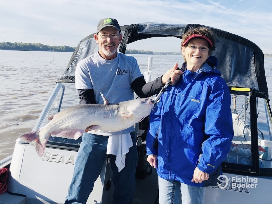 Two people are on a boat, and one is holding a large Catfish with a metal grip. They’re both smiling and dressed in outdoor clothing. There’s water and trees visible in the background.