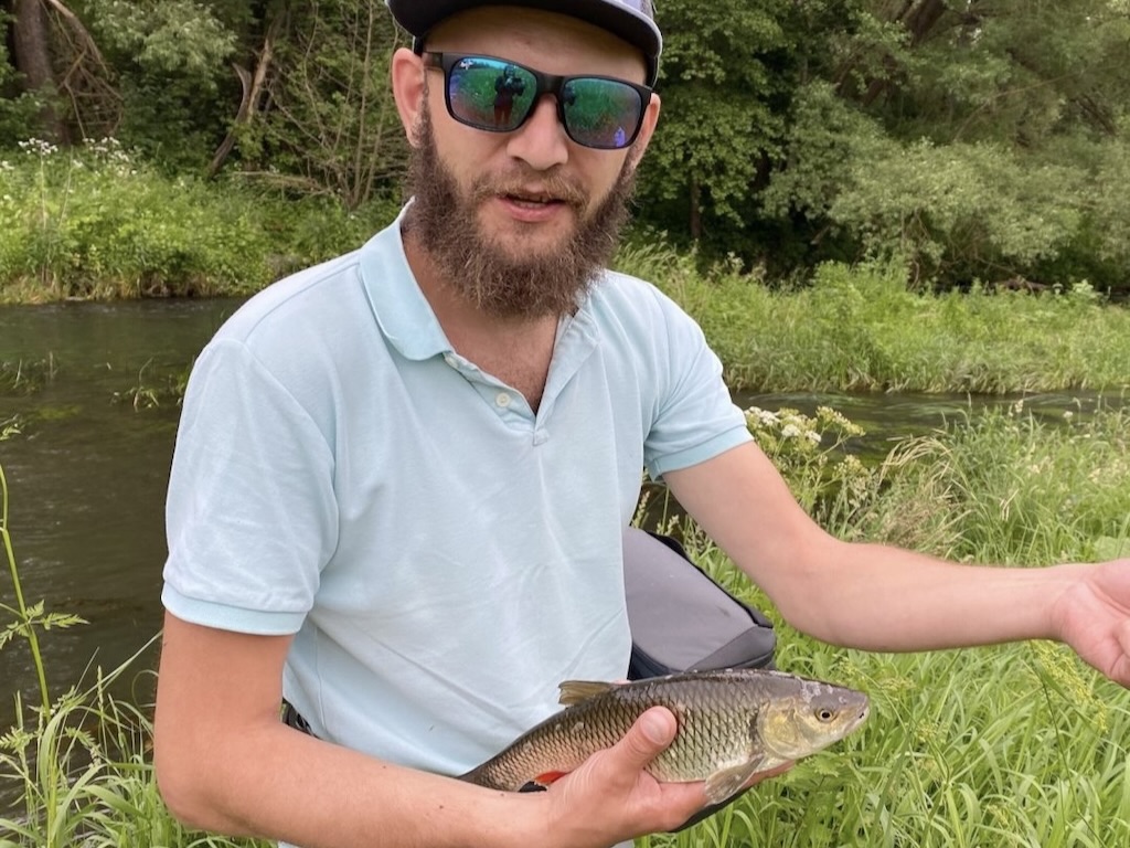 A man wearing sunglasses and a cap holds a Grass Carp near a riverbank with greenery. He’s been fishing for  on a sunny day.
