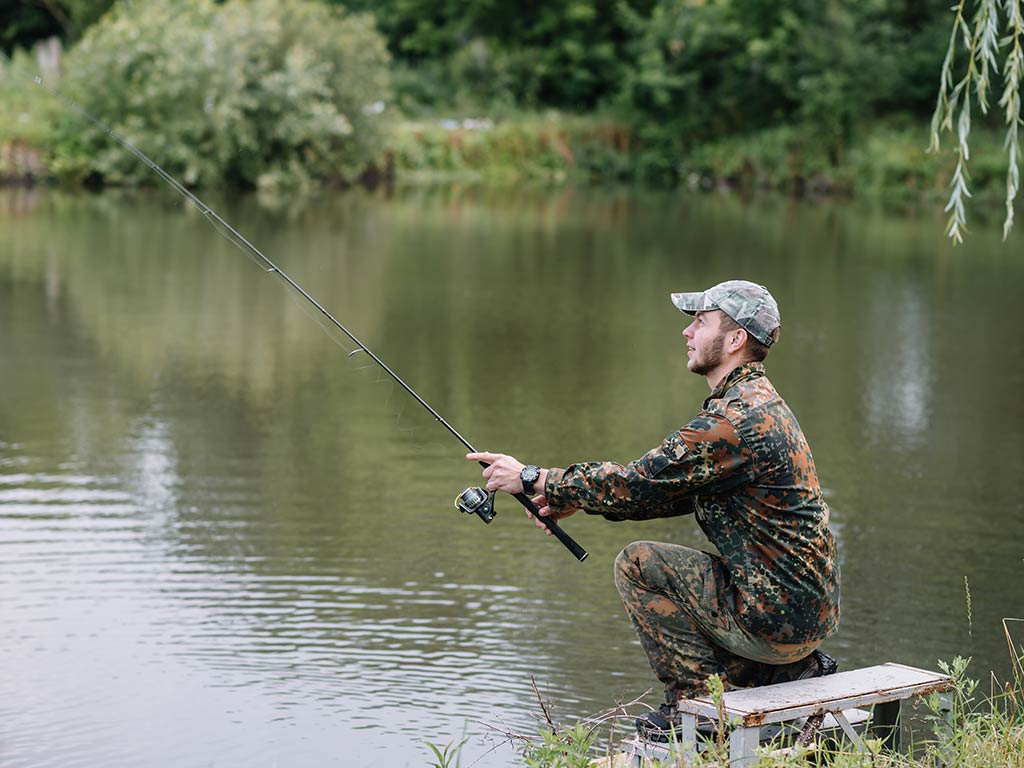 A man in camouflage clothing is sitting on a bench by a calm lake, fishing with his rod. He's surrounded by lush greenery, enjoying the peaceful atmosphere as he waits patiently for a bite.