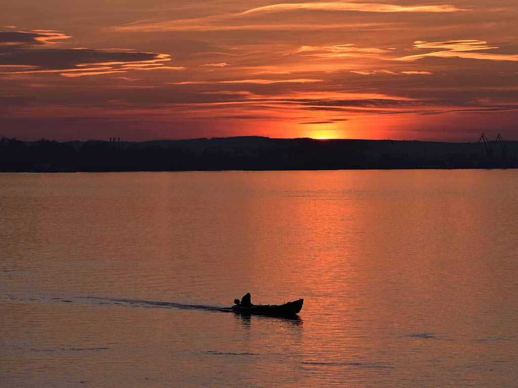 A view towards a silhouette of a person in a small boat rowing across calm water on the Danube River at sunset.
