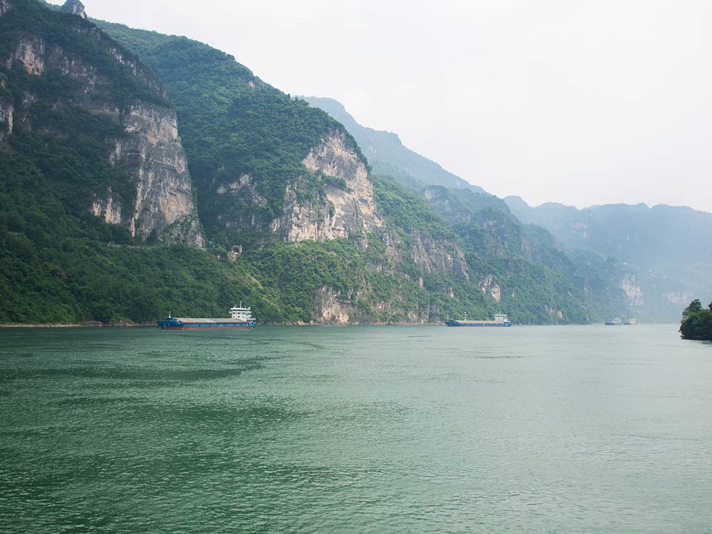 A wide river in China with calm water flows between steep, green mountains. Two cargo boats travel near the rocky shoreline, while locals are fishing under a cloudy sky.