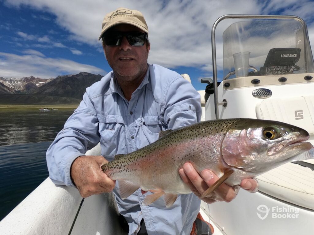 A person wearing sunglasses and a hat is holding a large Rainbow Trout on a boat, with mountains and a lake visible in the background.