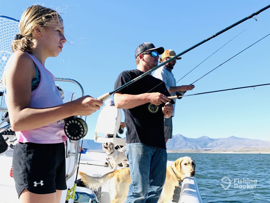 A girl and two men are standing on a boat, holding fishing rods, with two dogs nearby. In the background, you can see mountains and water, showing the scenery of Mammoth Lakes, CA.