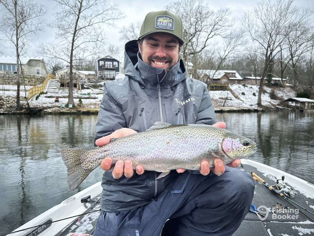 A man in a gray jacket and hat kneels on a boat and holds a Trout to the camera, with a snowy riverside backdrop.