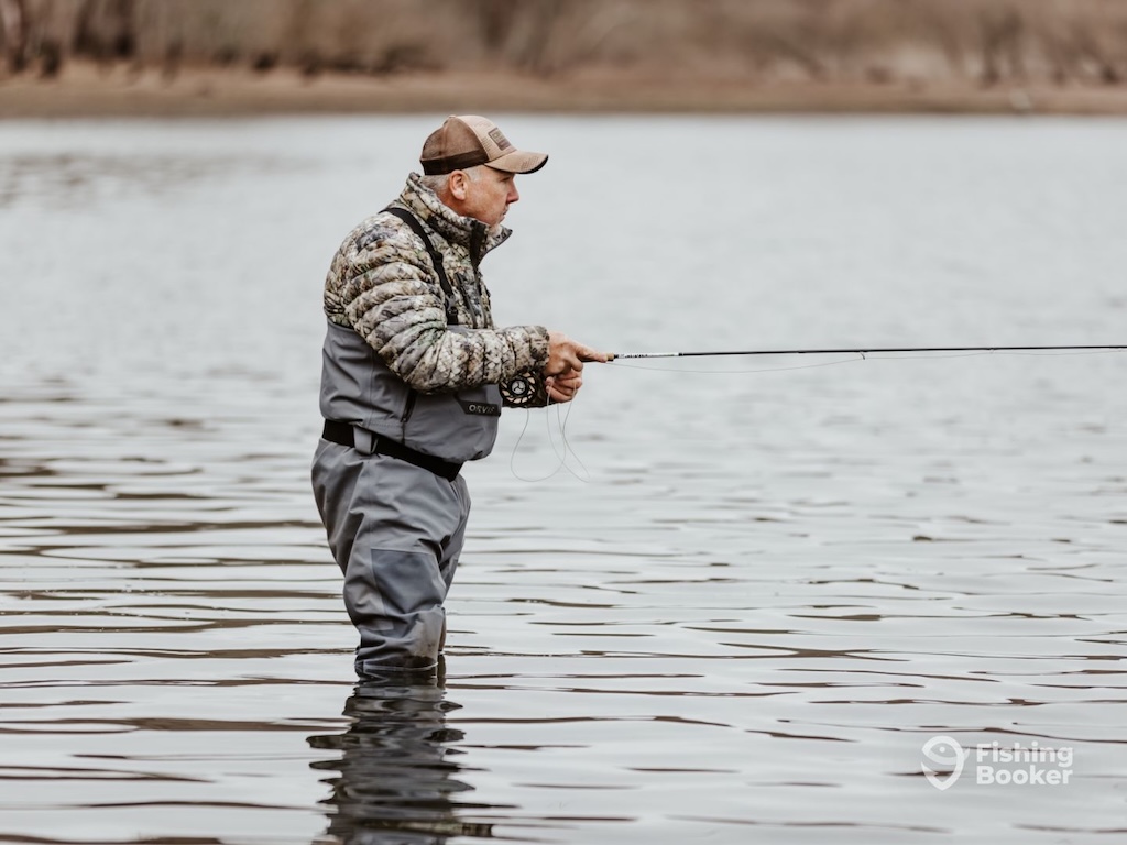 A man wearing waders and a cap stands in shallow water with a rod on an overcast day. 