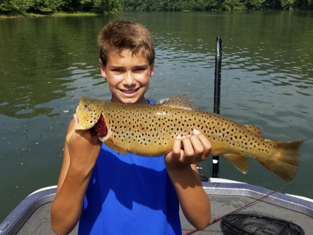 A boy in a blue tank top holding a large Brown Trout on a boat in Missouri with a scenic lake and trees in the background.