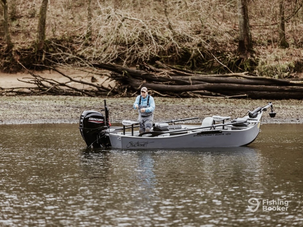 A person enjoys Trout fishing in Missouri, standing on a gray motorized boat in a calm river. Leafless trees and fallen branches line the shore.