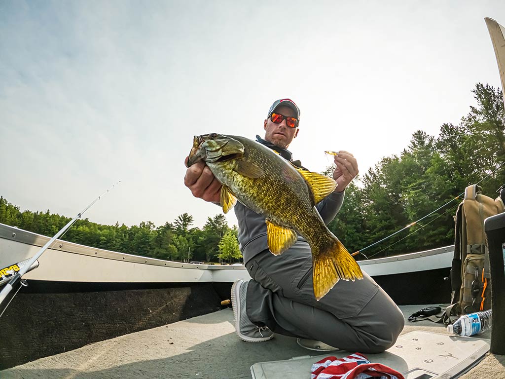 A person in sunglasses and outdoor gear is holding a large Smallmouth Bass and the lure that caught it while crouching on a boat, showing off their Minocqua fishing success. The background features trees and fishing equipment, adding to the outdoor atmosphere.